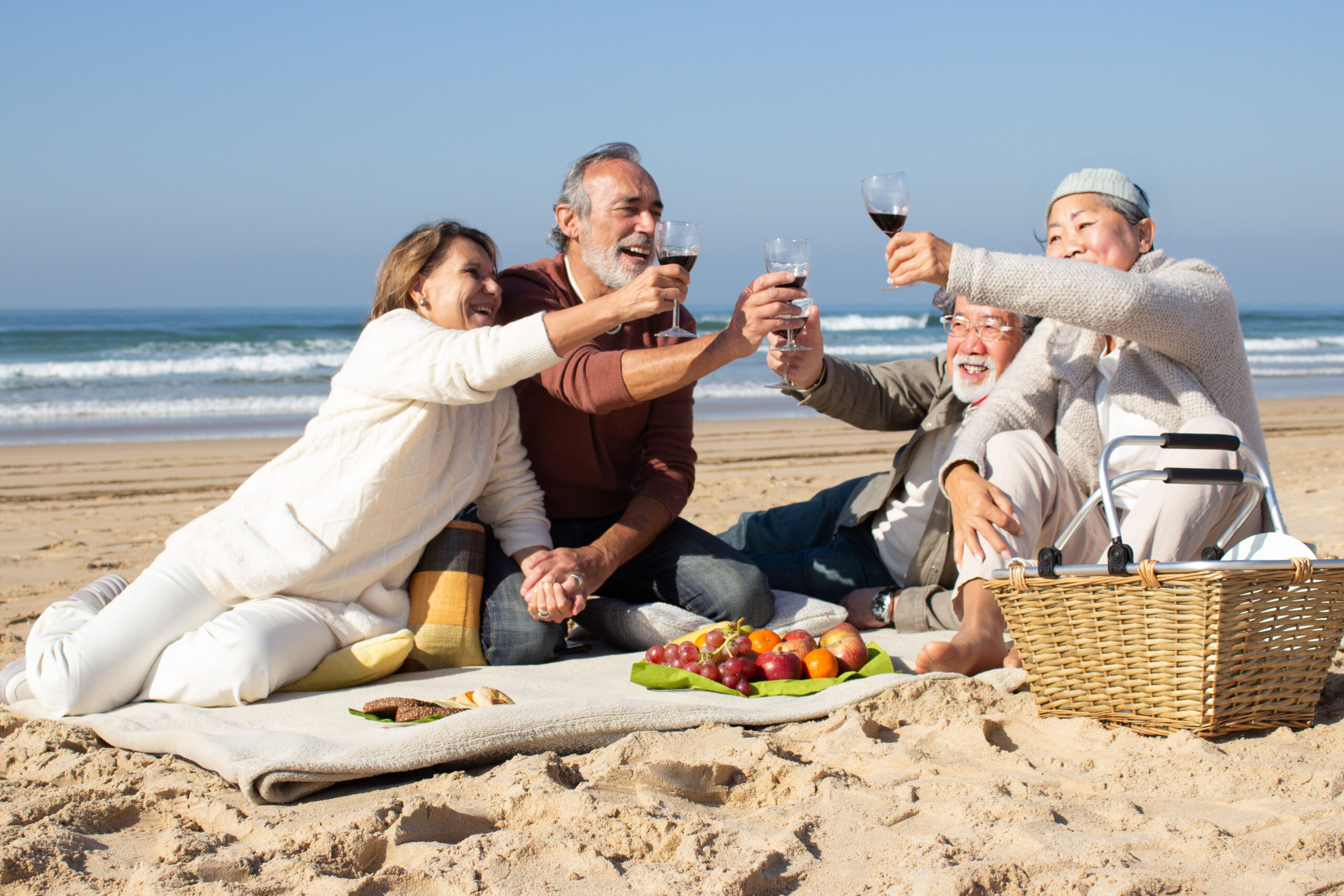 Four senior friends having picnic at beach on sunny day, raising glasses of red wine and pronouncing toast. Two middle-aged couples clinking glasses, celebrating outside. Celebration, leisure concept